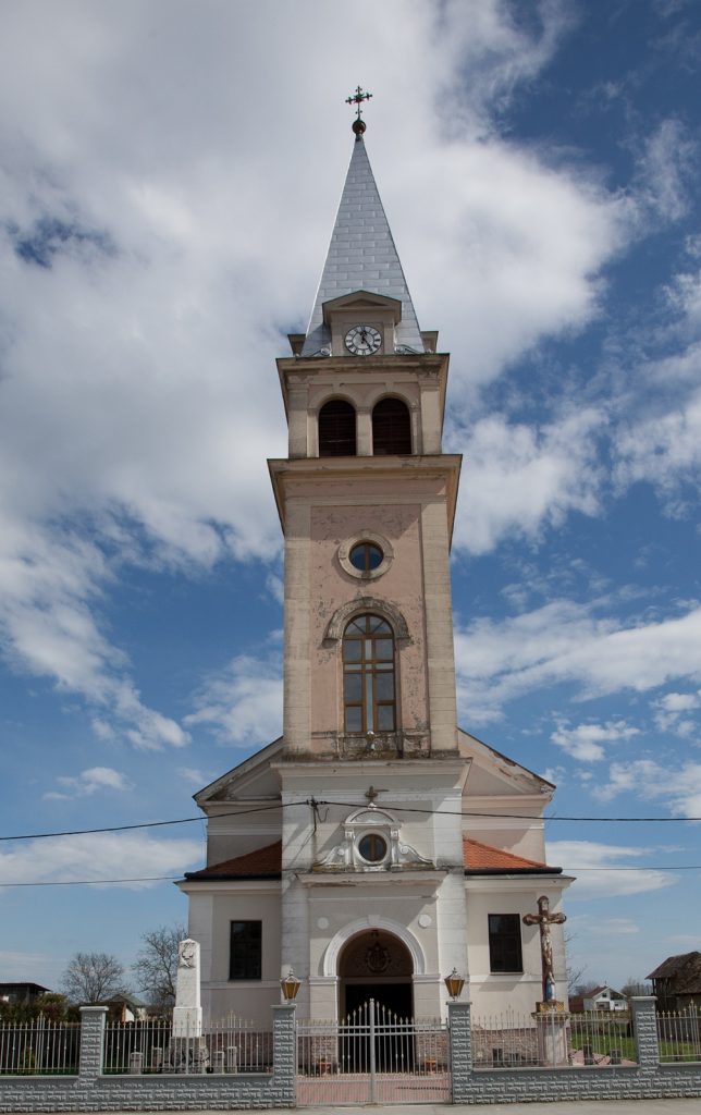 Tall church steeple with a clock, pointed silver roof, and cross atop against a blue sky with clouds.
