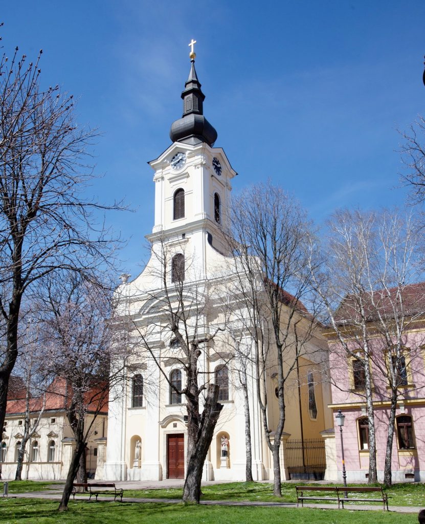 White church with a tall clock tower and dark onion dome, set against a blue sky with leafless trees in a park foreground.