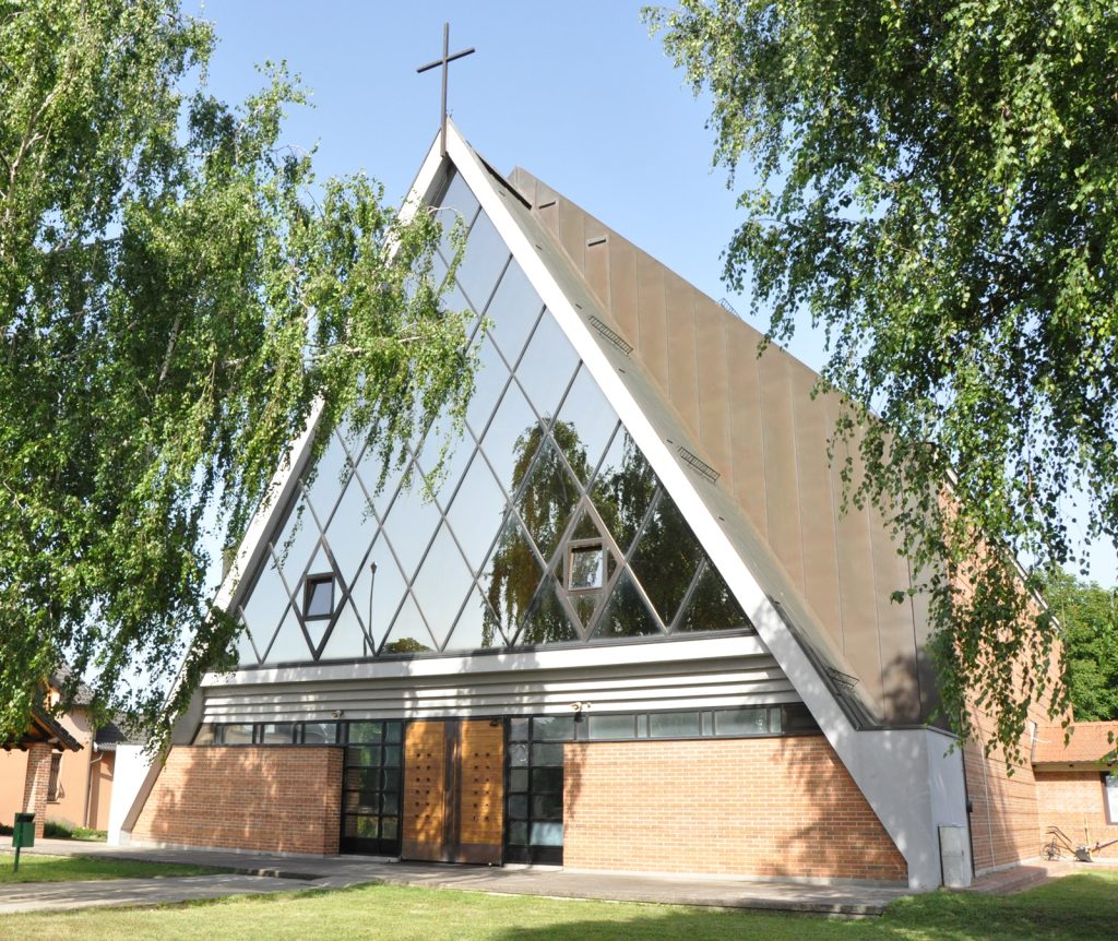 Modern church with a triangular glass facade and a cross on top, surrounded by green trees.