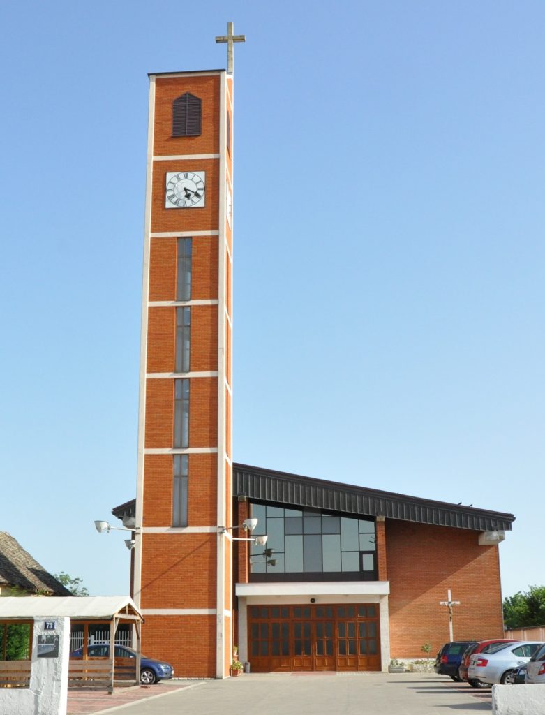 Brick clock tower with a cross atop, part of a brick church-like building against a clear blue sky.