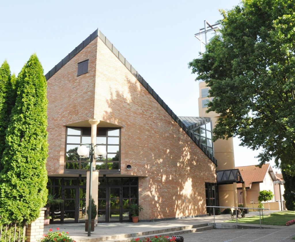 Brick building with a triangular roof and large glass entry, surrounded by trees and a street lamp in front.