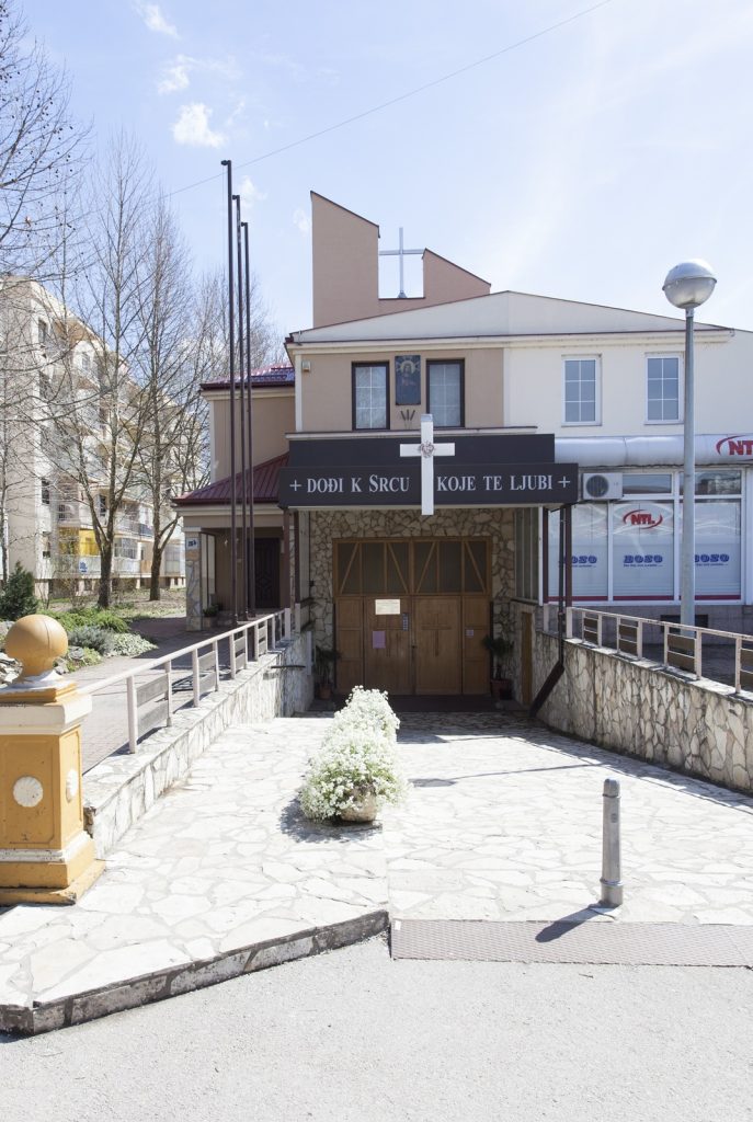 Front view of a small church entrance with a white cross on a black sign and wooden double doors, beige stone walls and a stone-paved walkway.