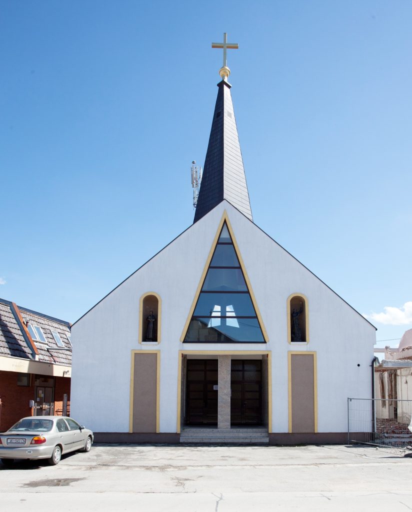 Modern church front with triangular facade, central glass window, and cross-topped steeple against a blue sky.