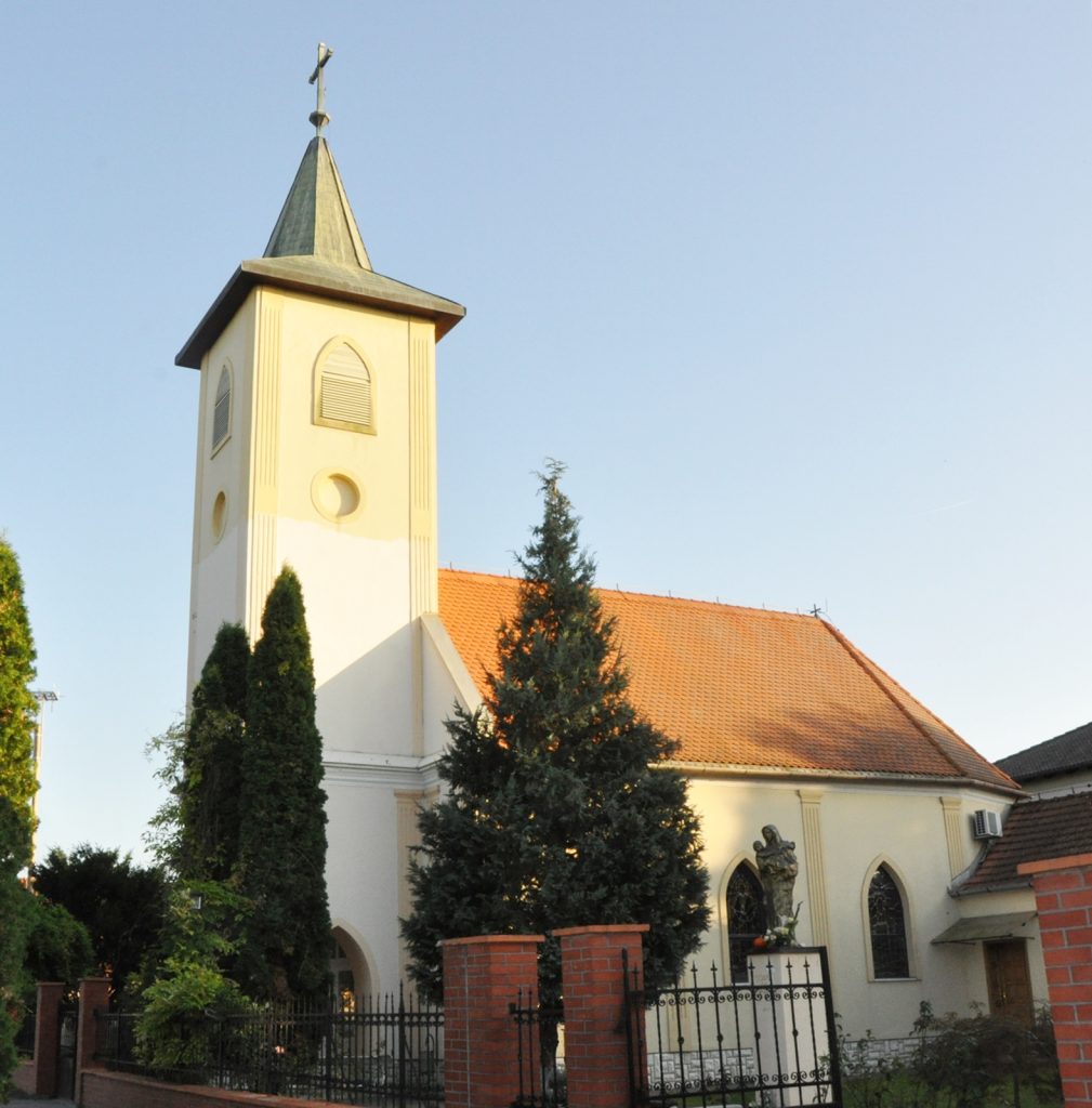 Church with a tall beige steeple and cross atop, orange-tiled roof, and gated yard with evergreens nearby.