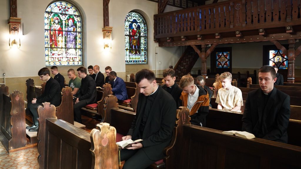 Group of men in church pews, some in clerical collars, praying with Bibles open before them near stained-glass windows and wooden decor.