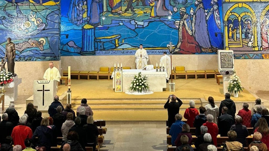 Church interior during Mass with an altar, two priests, and a congregation, set against a large colorful mural.