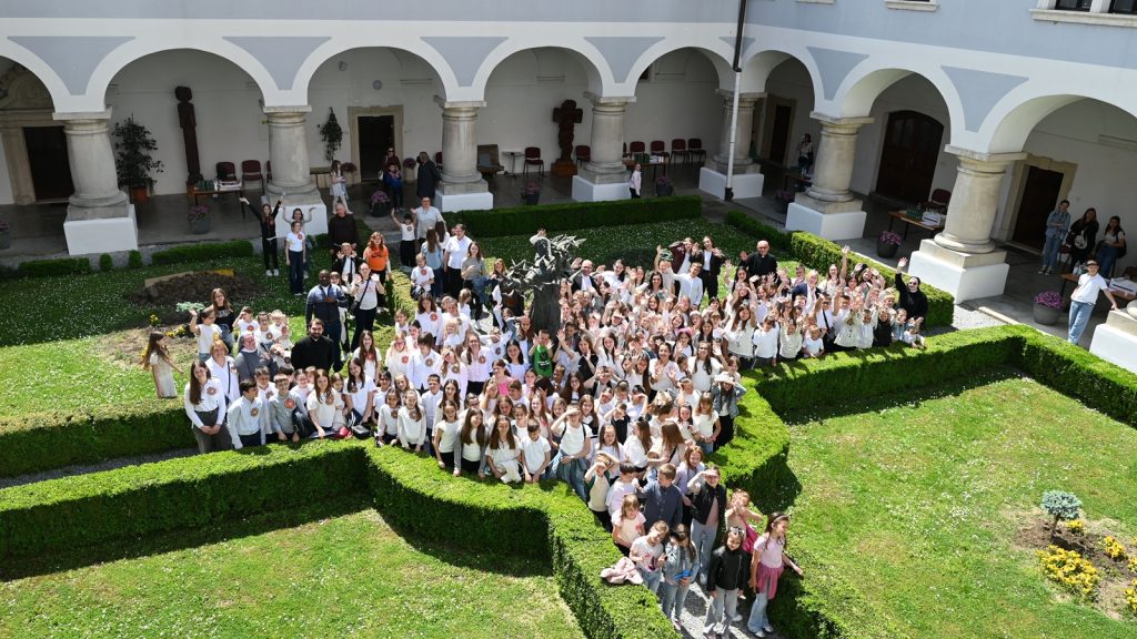 Large group of children and adults in white shirts pose for a group photo in a courtyard with arched colonnades and trimmed hedges surrounding a central garden bed.