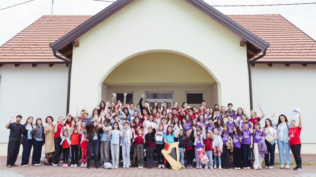 Large group of children and adults waving from the steps of a pale building with an arch entrance and red-tiled roof.