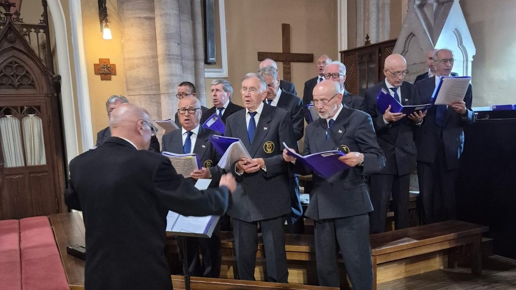 Conductor faces a choir of elderly men in gray suits singing from purple folders inside a church.