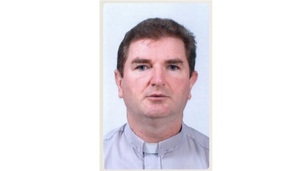 Head-and-shoulders portrait of a Catholic priest wearing a clerical collar. 16:9 frame with light background.