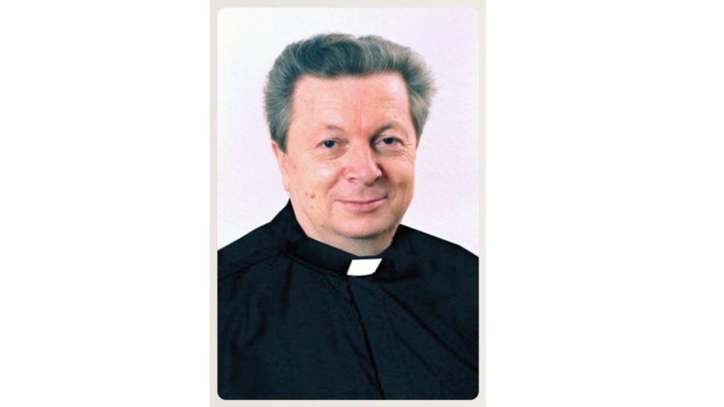 Portrait of a smiling Catholic priest in a black cassock with a white clerical collar, looking at the camera.
