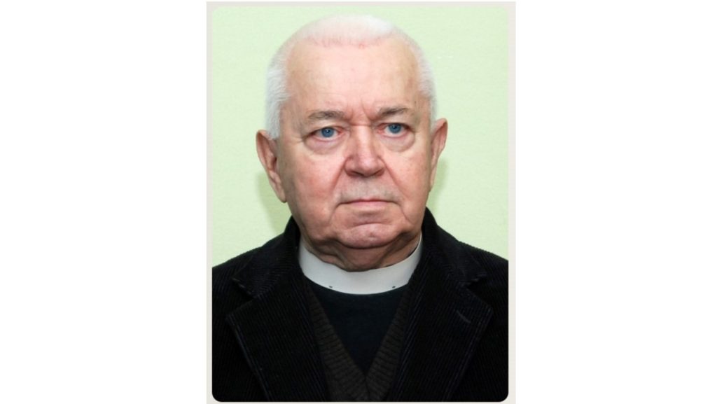 Portrait of an elderly man with white hair and blue eyes, wearing a black jacket and a clerical collar, looking straight ahead.
