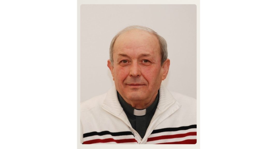 Portrait of an older man with a clerical collar, wearing a white sweater with red and black stripes, facing the camera.