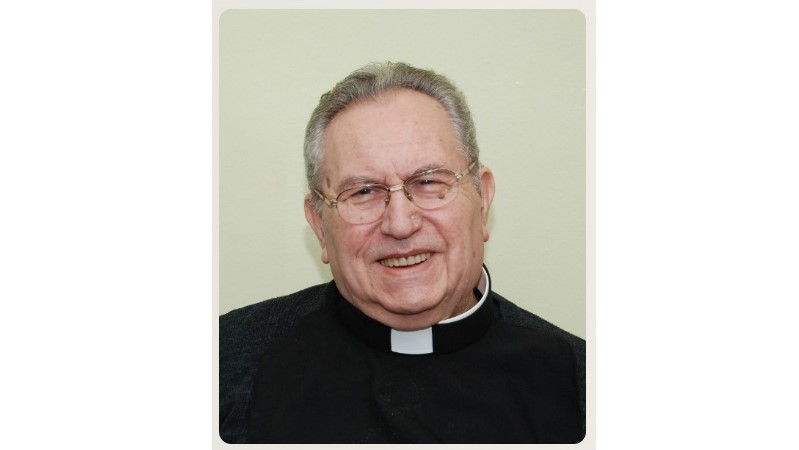 Portrait of an older priest smiling, wearing glasses and a black clerical shirt with a white collar.