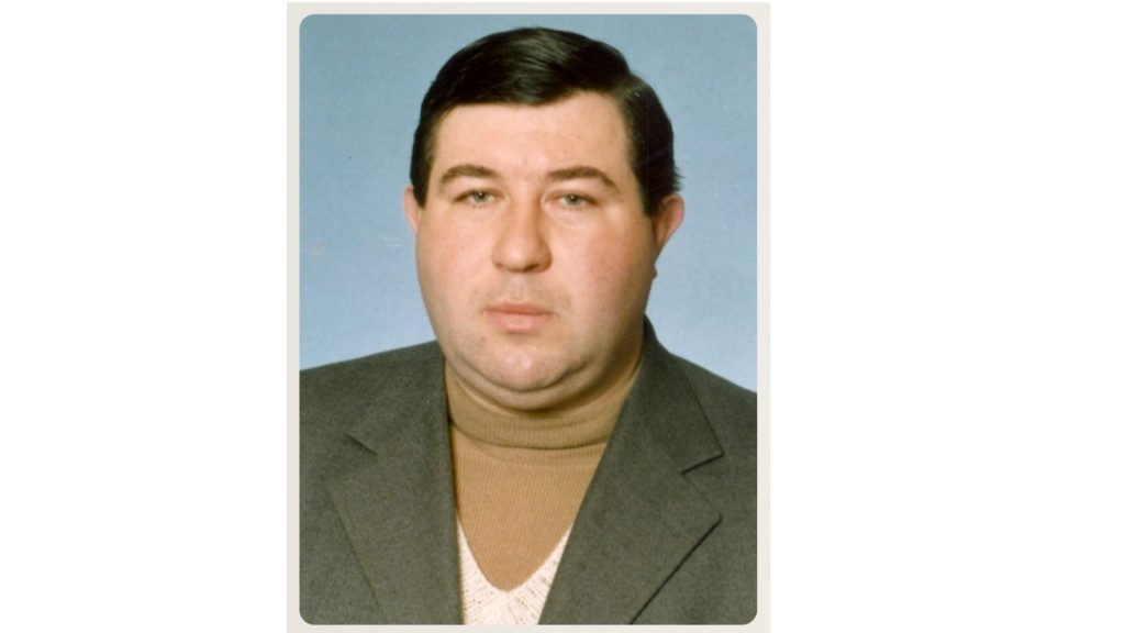 Portrait of a middle-aged man with dark hair in a brown turtleneck and gray blazer against a blue backdrop.