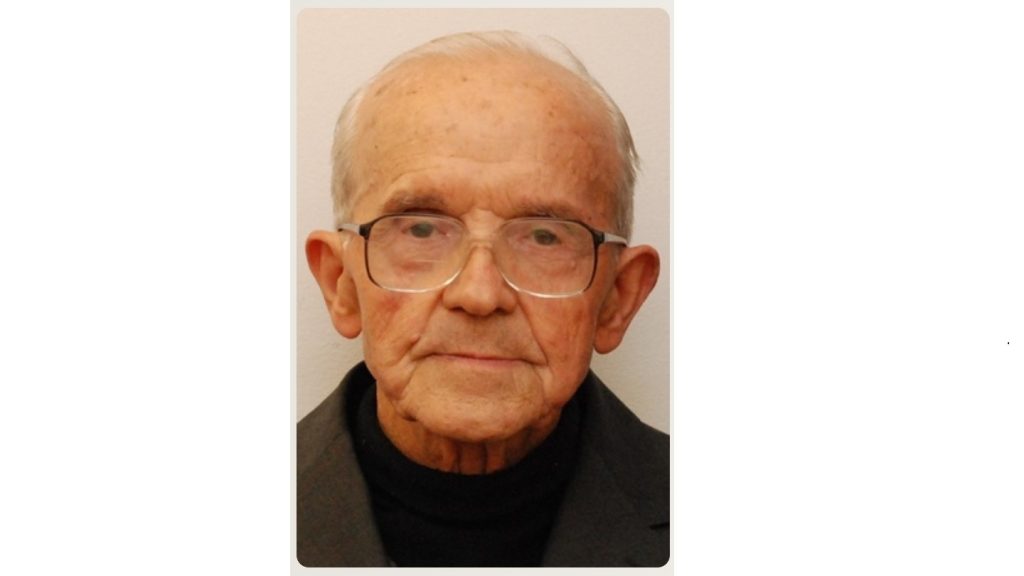 Portrait of an elderly man with gray hair and glasses, wearing a dark jacket, looking at the camera against a light background.