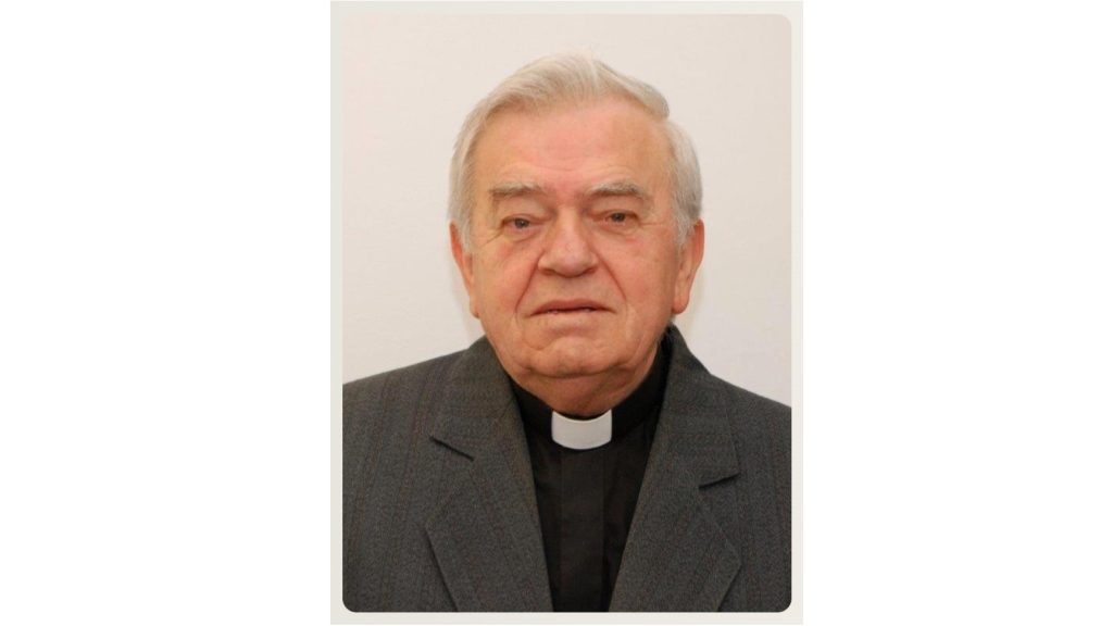 Portrait of an elderly priest wearing a black clerical collar and gray suit, facing the camera with a neutral expression.