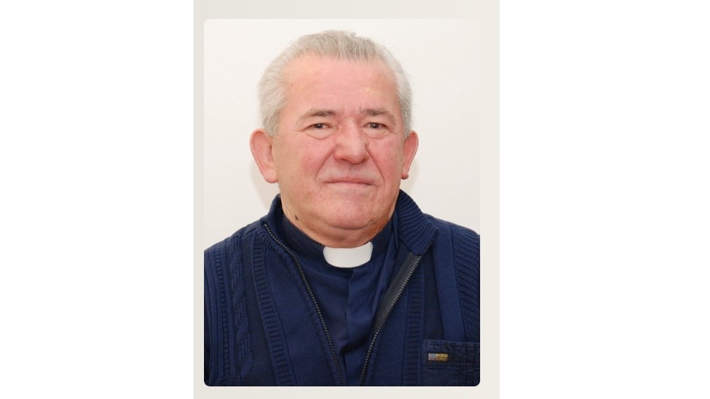 Older man wearing a clerical collar and navy sweater, looking at the camera.