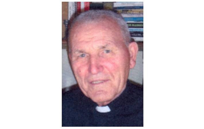 Older man with short gray hair wearing a clerical collar, looking at the camera in a home setting.