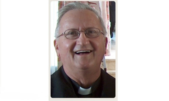 Smiling Catholic priest in a black shirt and white clerical collar indoors.
