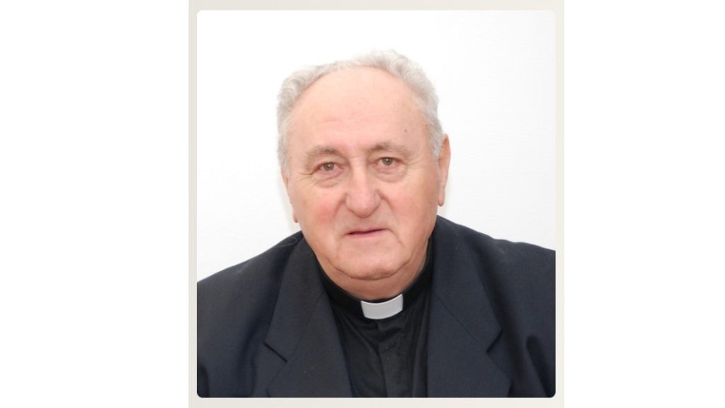 Portrait of a senior priest in a black clerical shirt and white collar, facing the camera against a light background.