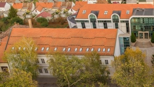 Aerial view of orange-tiled buildings with white facades and a modern glass-fronted structure among trees.