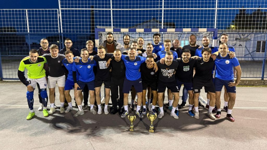 Group of men in soccer/futsal gear posing together on an outdoor court at night with two trophies in front.
