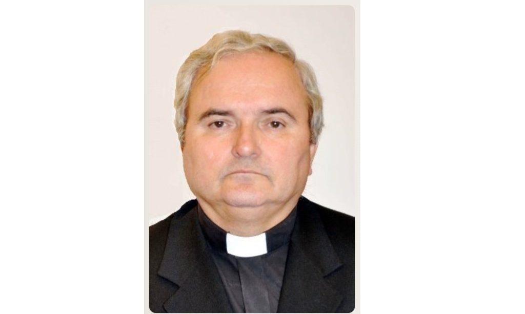 Portrait of a middle-aged man wearing a black suit and white clerical collar, facing the camera against a light background.