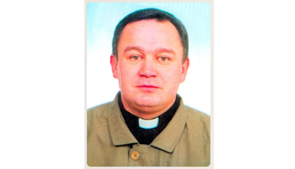 Head-and-shoulders portrait of a man wearing a clerical collar and tan shirt, looking at the camera.