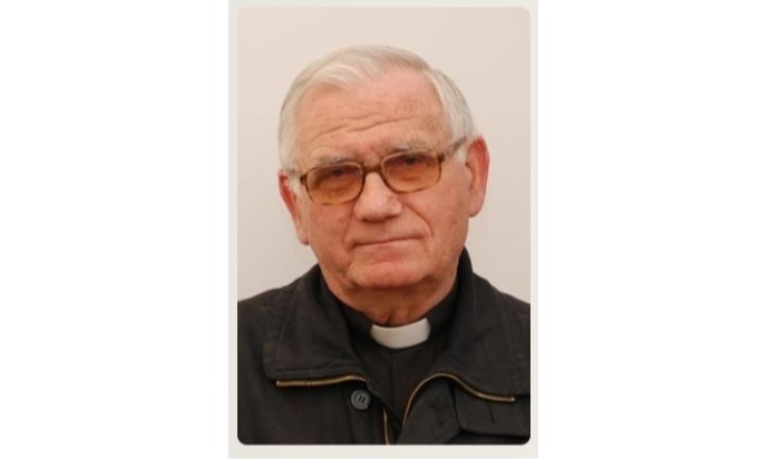 Older man with white hair and glasses, wearing a clerical collar and dark jacket, looking at the camera.
