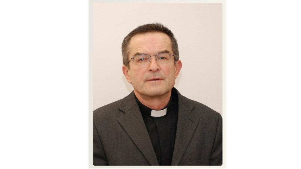 Portrait of a middle-aged priest in a gray suit and clerical collar facing the camera.