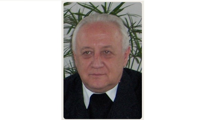 Portrait of an older man with white hair wearing a suit and tie, indoors with green plants in the background, looking at the camera.
