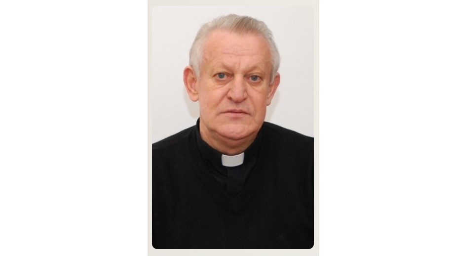 Portrait of an older man with short gray hair wearing a black clerical shirt and white collar, looking at the camera.
