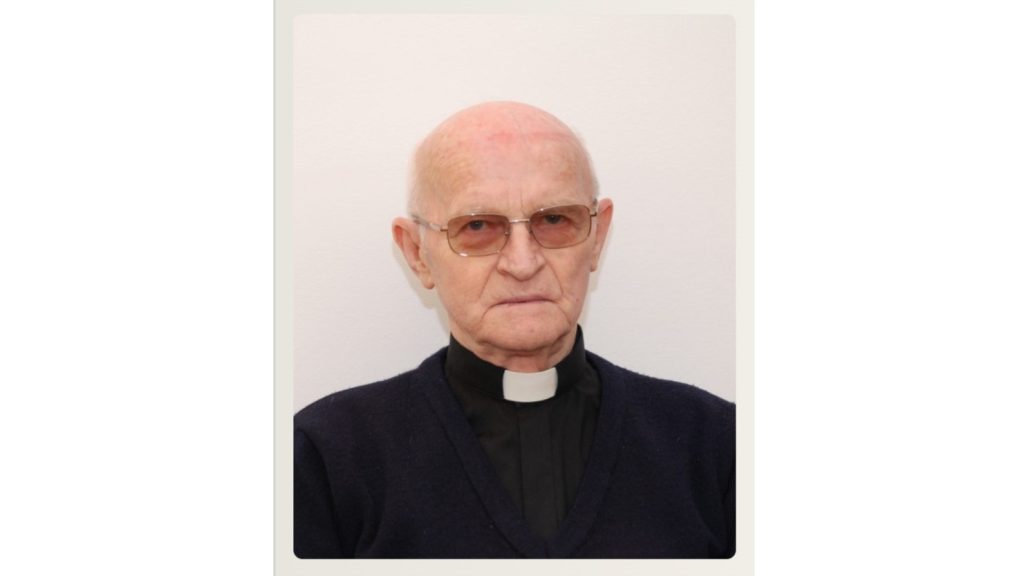 Portrait of an older man in a clerical collar and dark sweater, looking at the camera against a neutral background with a light wall behind him.