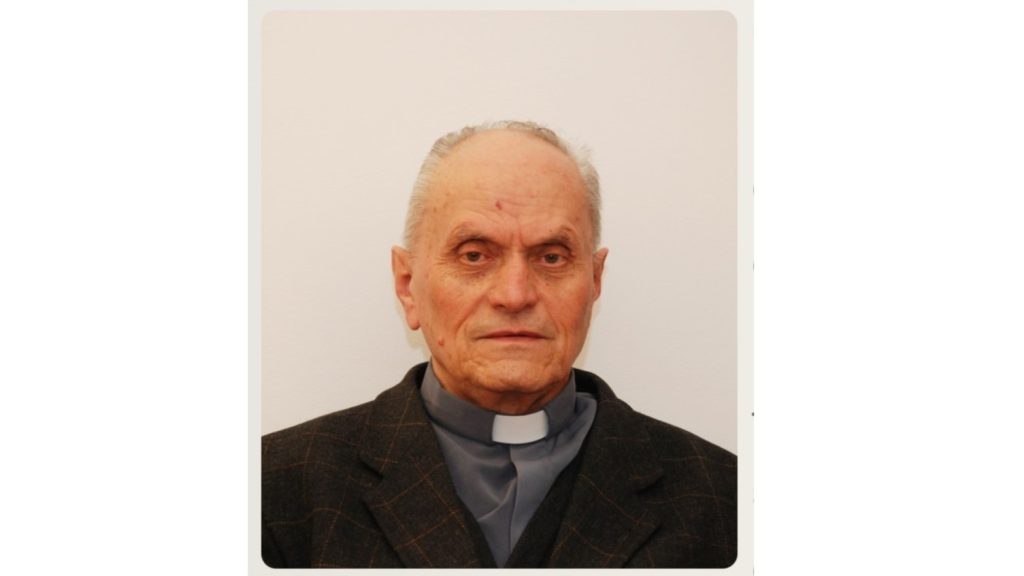 Portrait of an older man with a clerical collar and dark blazer against a light background, looking straight ahead.