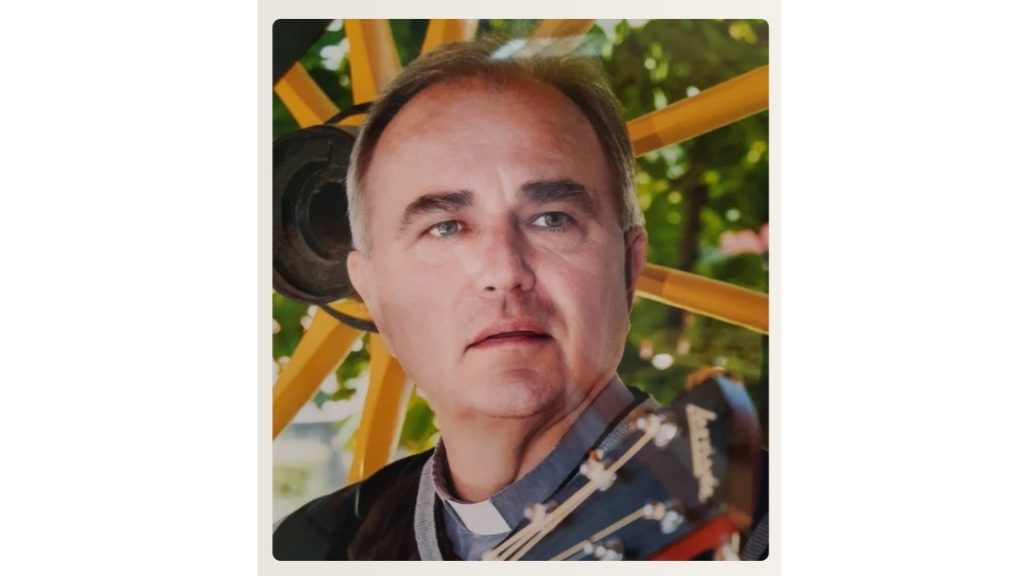 Portrait of a middle-aged man wearing a clerical collar, looking to the side, with a guitar neck in the foreground and colorful outdoor equipment behind him.