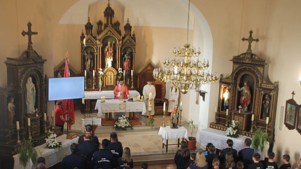 Inside a church during mass, a priest at a decorated altar with candles and statues, facing an attentive congregation.