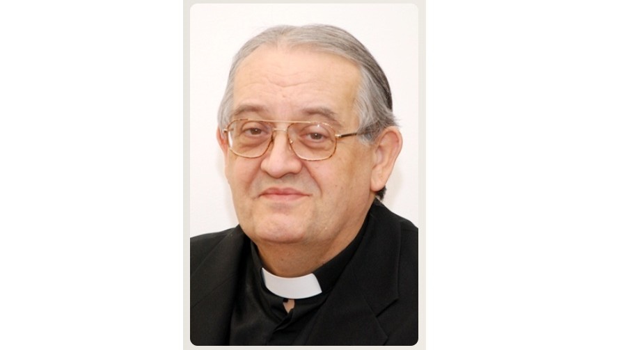 Portrait of an older Catholic priest wearing glasses and a black clerical shirt with a white collar, smiling slightly.