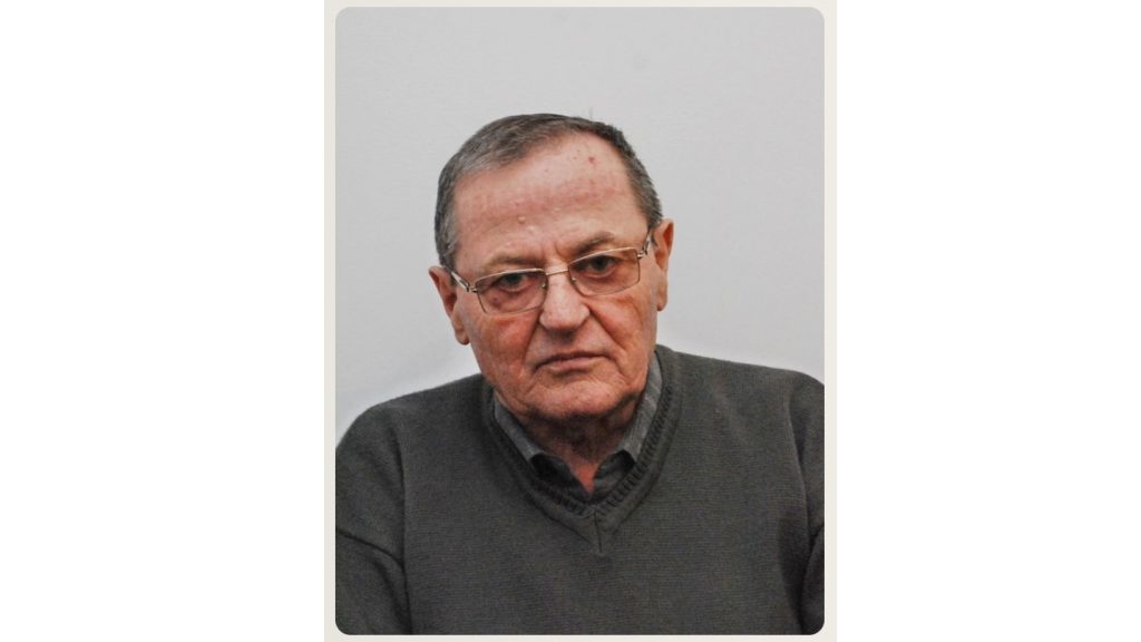 Portrait of an older man with short gray hair and glasses, wearing a dark sweater, looking slightly to the left against a pale background.