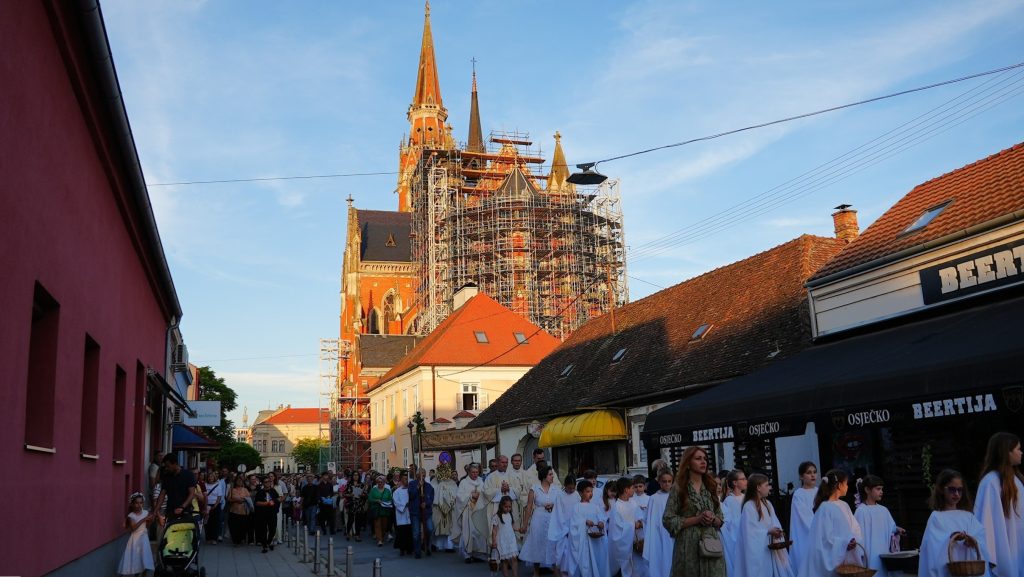 Religious procession of people in white robes walking down a sunlit street with a Gothic cathedral under scaffolding in the background.