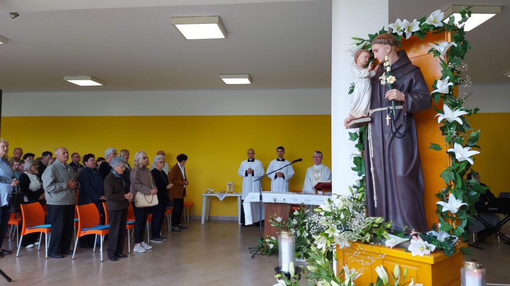 Congregation standing in a bright room with a yellow wall, facing an altar where priests in white vestments lead a service; a large statue of a saint surrounded by white lilies is arranged on the right.