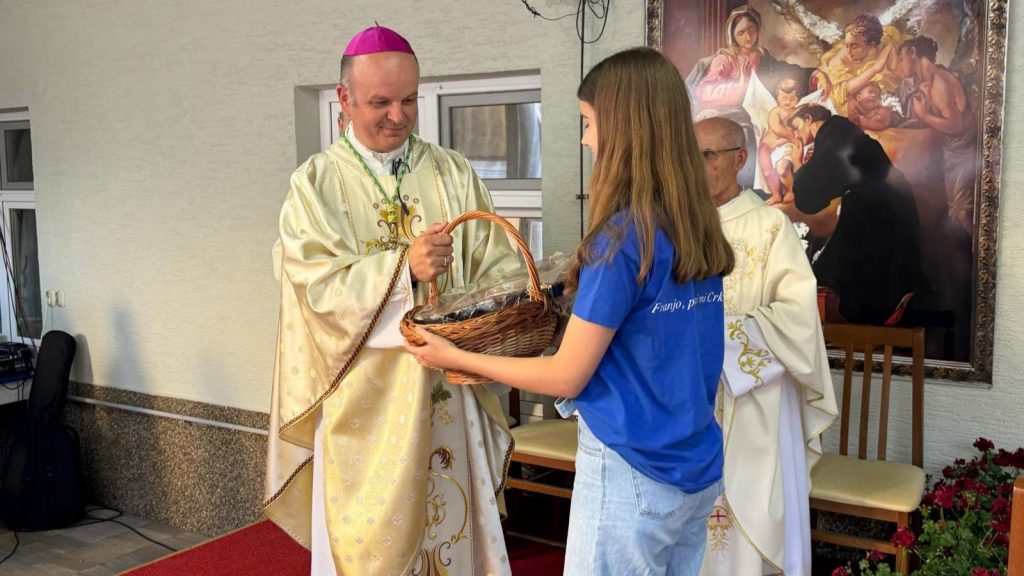 Catholic bishop in cream vestments hands a wicker gift basket to a young woman in a blue shirt during a ceremony outside a church.