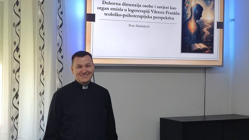 Smiling priest in a black clerical shirt standing beside a projection screen in a conference room with patterned curtains on the left.