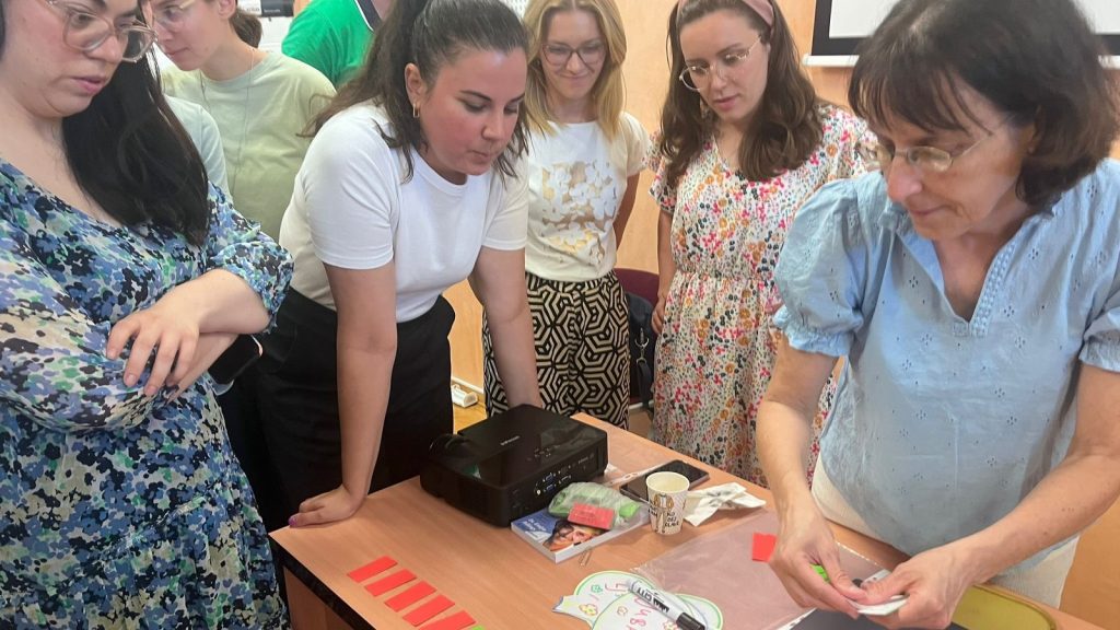 Group of women gathered around a table watching an instructor fold paper during a crafting workshop.