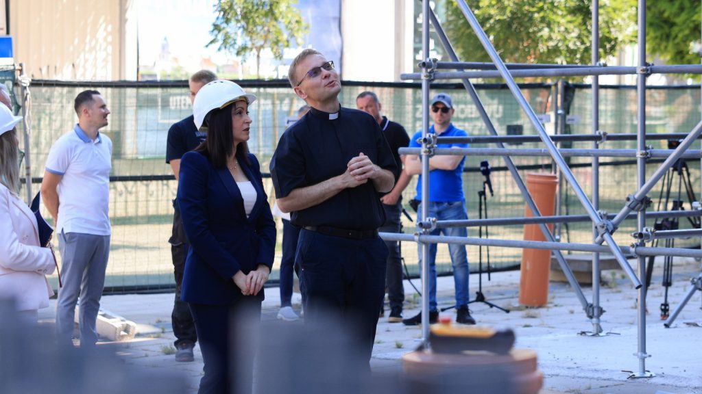 Priest and a woman in a hard hat listen to a briefing at a construction site with scaffolding behind them.