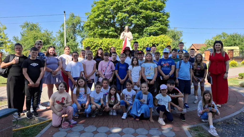 Large group of children posing for a outdoor group photo in a park with a statue in the background. Two adults stand at edges, sunny day, everyone looking at the camera.