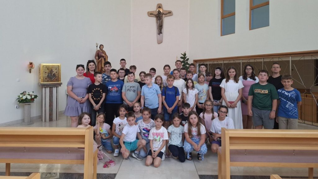 Group of children and teens posing for a photo inside a church, standing in front of a crucifix and altar area.