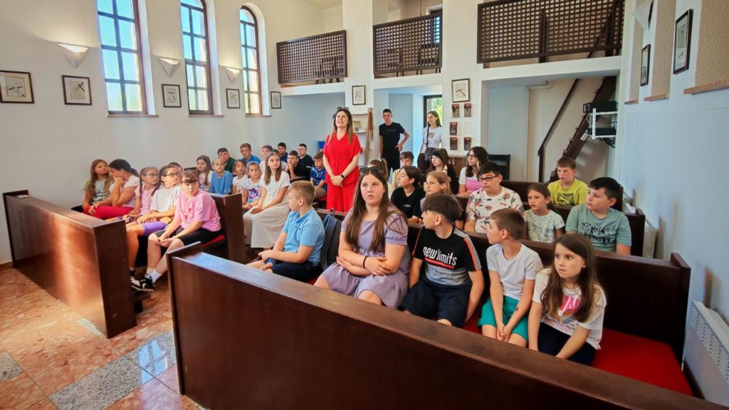 Group of children seated on wooden pews in a bright hall as a woman in a red dress stands at the front, likely leading a session in a church-like room.