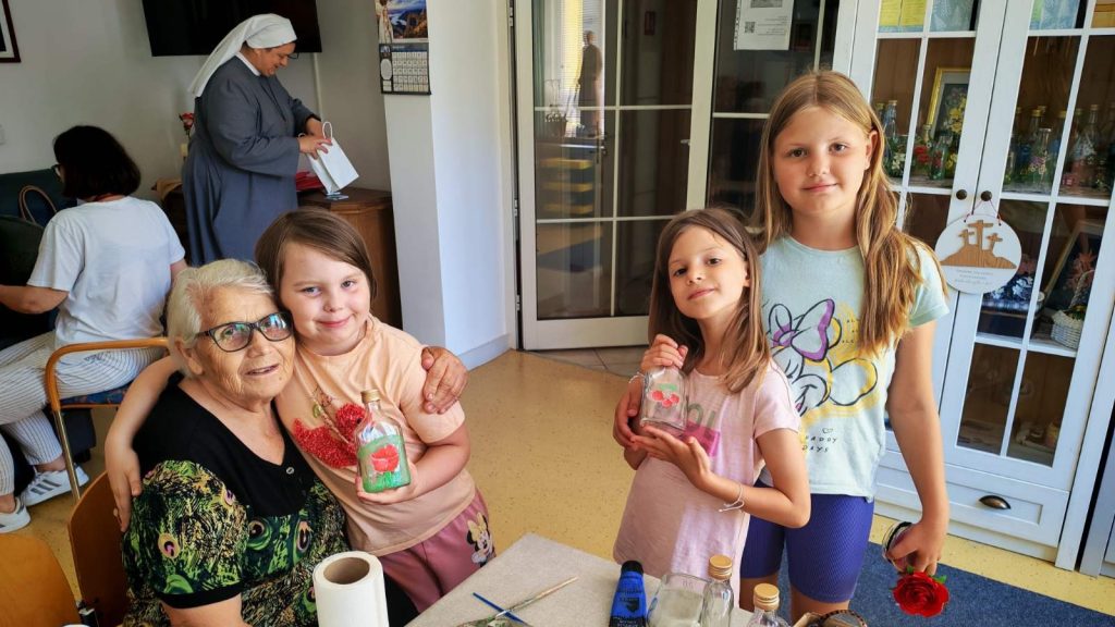Older woman with white hair and glasses sits with two smiling girls, who hold decorated painted bottles in a craft room.