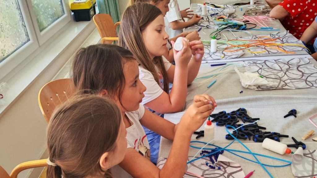 Children sit at a long table crafting with beads, strings, and glue in a bright room.